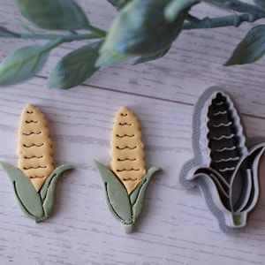 May include: Two corn-shaped biscuits and a corn-shaped biscuit cutter. The biscuits have a light yellow centre and green husks. The cutter is grey and white. The items are on a light wood surface.