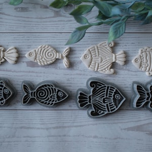 Puede incluir: Vista cenital de seis cortadores de galletas con forma de pez. La fila superior presenta cuatro cortadores blanquecinos con diseños detallados. La fila inferior muestra dos cortadores negros y grises con patrones intrincados. Fondo de madera clara.