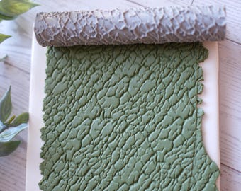 May include: A textured rolling pin rests above a green, textured surface. The rolling pin has a gray, bark-like pattern. The green surface has a repeating, organic pattern. The items are on a white surface.