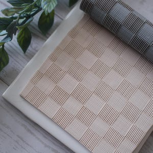 May include: A close-up shot of a beige and gray woven placemat and a rolled-up gray woven mat. The mats have a square pattern. A sprig of green leaves is in the upper left corner.