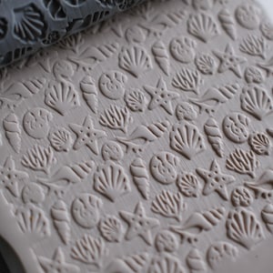 May include: A gray rolling pin with a seashell pattern is pressed into a beige clay surface. The embossed design includes various seashells and starfish, creating a textured, beach-themed pattern. The clay is ready for crafting.