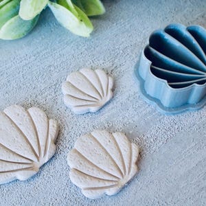 以下が含まれることがあります： 水色の貝殻型クッキーカッターと、3つの貝殻型クッキー。クッキーはベージュ色で、詳細なリブがあります。クッキーカッターはプラスチック製です。背景は明るい灰色のテクスチャ加工された表面です。