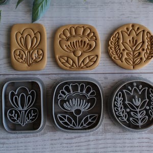 May include: Six floral-shaped cookie cutters and their corresponding embossed cookies. The cutters are silver-colored, and the cookies are light brown. The designs include flowers and leaves in rectangular and circular shapes.