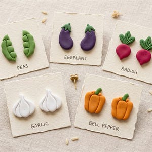 Könnte beinhalten: Handgefertigte Ohrstecker mit Gemüse-Motiven. Die Ohrringe zeigen Erbsen, Auberginen, Radieschen, Knoblauch und Paprika. Jedes Paar wird auf einer strukturierten Karte mit dem Namen des Gemüses präsentiert. Die Ohrringe sind farbenfroh und skurril.