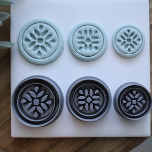 May include: Six round cookie cutters in two colors, arranged in two rows. The top row features three light blue cutters, while the bottom row has three dark gray cutters. Each cutter has a floral design.