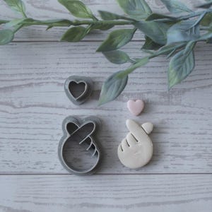 Puede incluir: Cortadores de galletas en forma de corazón y de mano, junto con un pequeño corazón rosa. Los cortadores son grises y blancos, colocados sobre una superficie de madera clara. Se ven ramitas verdes en el fondo.