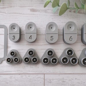 May include: A collection of gray plastic tools, including a rectangular frame with "1.5MM" text, and several oval and triangular pieces with numbers 3 through 7. The tools have small holes and are arranged on a light wood surface.