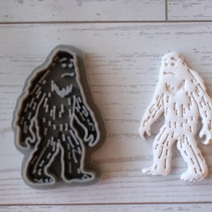 Puede incluir: Un cortador de galletas con forma de Bigfoot junto a una galleta con forma de Bigfoot. El cortador es de color gris oscuro y la galleta es blanca. Ambos tienen una expresión triste y una apariencia peluda, con líneas de pelaje detalladas.