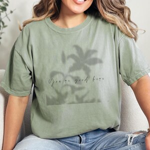 May include: Sage green t-shirt with a palm tree silhouette design and the text "You're good here." The shirt is made of a soft material and is paired with blue jeans. The model is wearing a gold bracelet.