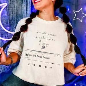 以下が含まれることがあります： ロシア語と英語で「I love you」と書かれたクリーム色のTシャツ。Tシャツには、「All The Things She Said」というテキストが入った音楽プレーヤーのグラフィックが描かれています。