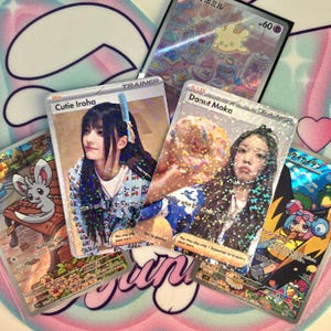 Puede incluir: Varias cartas coleccionables holográficas con ilustraciones de estilo anime y retratos de mujeres jóvenes. Las cartas tienen texto en japonés e inglés, incluyendo los nombres "Cutie Iroha" y "Donut Moka". Las cartas están sobre una superficie con estampado rosa y blanco.
