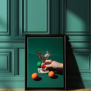 Puede incluir: Una impresión enmarcada que presenta una copa de martini sostenida por una mano con esmalte de uñas rojo, descansando sobre un billete de dólar. Dos esferas naranjas y una verde están sobre una superficie verde. El fondo es una pared verde azulado.
