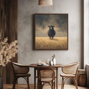 K&ouml;nnte beinhalten: Ein gerahmtes Kunstwerk zeigt ein schwarzes Schaf, das in einem Feld aus hohem, goldenem Gras unter einem st&uuml;rmischen Himmel steht. Das Kunstwerk h&auml;ngt &uuml;ber einem Holzesstisch mit Korbst&uuml;hlen und schafft ein rustikales und gem&uuml;tliches Interieur.