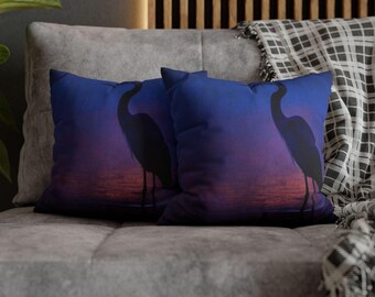 May include: Two square throw pillows with a vibrant sunset scene. The pillows feature a silhouette of a heron against a gradient of blue, purple, and orange hues. The pillows are placed on a gray couch, with a patterned blanket in the background.