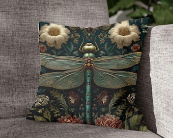 Puede incluir: Cojín cuadrado decorativo con un diseño detallado de libélula. El cojín tiene un fondo verde oscuro con elementos florales y botánicos. La libélula es verde azulado y dorada, con alas grandes y un cuerpo detallado. El cojín está sobre un sofá gris.