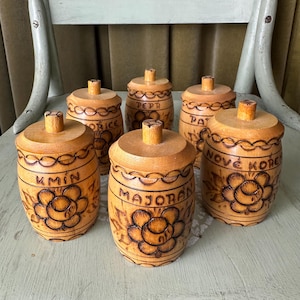 May include: A set of six small, light-brown wooden spice containers with lids, each featuring a barrel-like shape. The containers are decorated with wood-burned floral patterns and labels including text such as KMIN, MAJORAN, and NOVÉ KOREN.