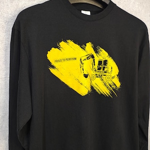 May include: Black long-sleeve shirt with a yellow graphic of a construction vehicle and the text "PROUD TO PERFORM". The graphic is on a yellow brushstroke background. The shirt is hanging on a black hanger.