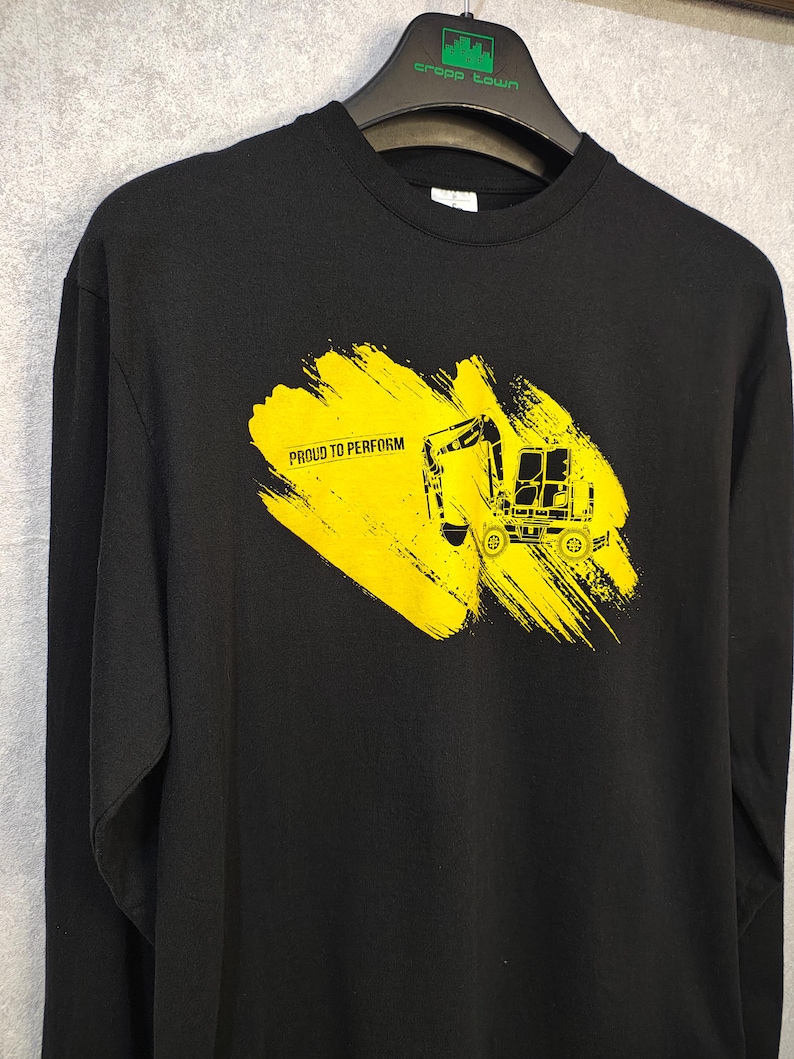 May include: Black long-sleeve shirt with a yellow graphic of a construction vehicle and the text "PROUD TO PERFORM". The graphic is on a yellow brushstroke background. The shirt is hanging on a black hanger.