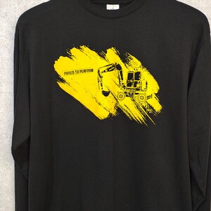 May include: Black long-sleeve sweatshirt featuring a yellow excavator graphic on a yellow brushstroke background. The text "PROUD TO PERFORM" is printed below the excavator. The sweatshirt is hanging on a black hanger.