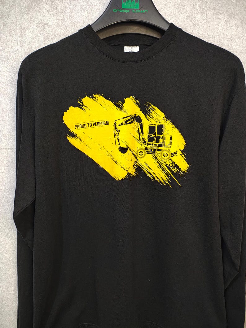 May include: Black long-sleeve sweatshirt featuring a yellow excavator graphic on a yellow brushstroke background. The text "PROUD TO PERFORM" is printed below the excavator. The sweatshirt is hanging on a black hanger.