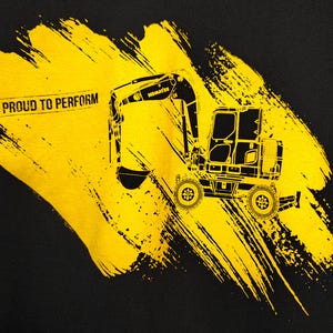 May include: Black t-shirt featuring a yellow excavator graphic with the text "PROUD TO PERFORM". The excavator is a detailed illustration with the brand name "KOMATSU" visible. The design includes yellow brushstroke accents.
