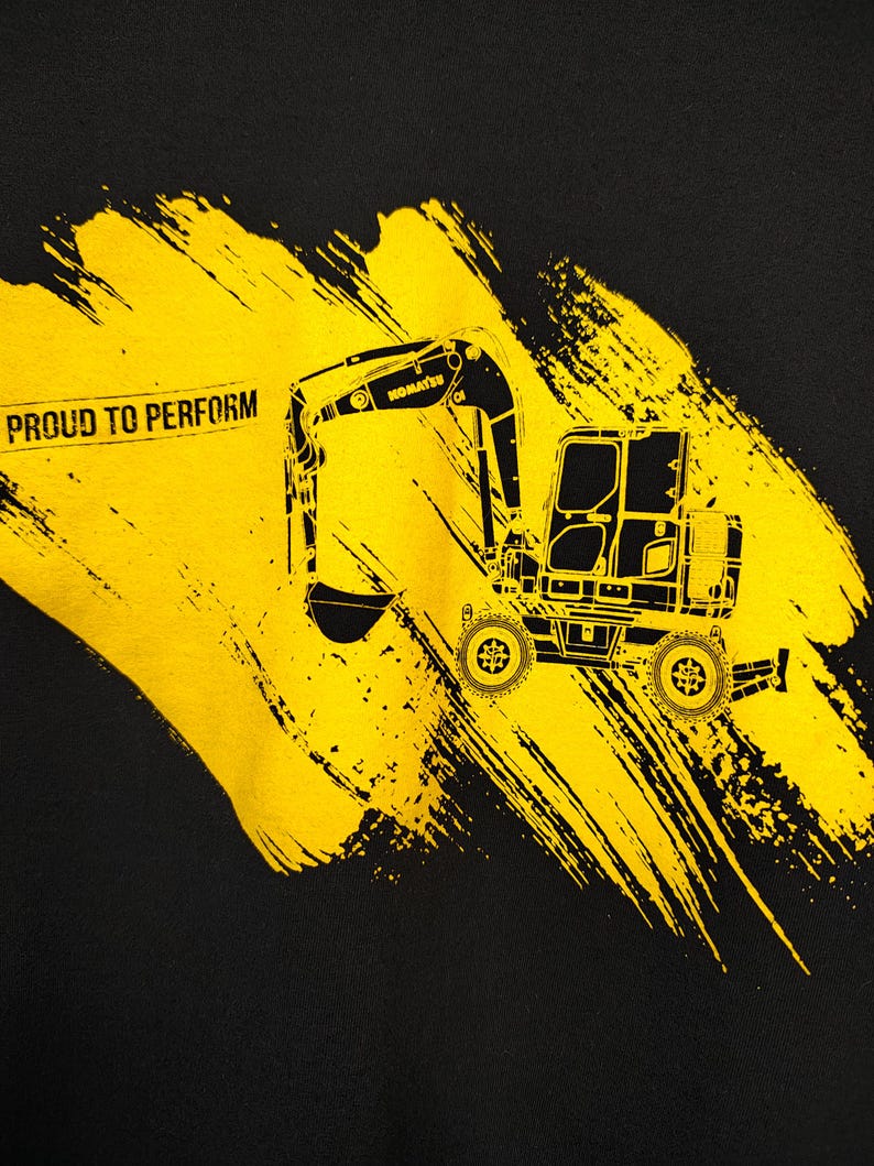 May include: Black t-shirt featuring a yellow excavator graphic with the text "PROUD TO PERFORM". The excavator is a detailed illustration with the brand name "KOMATSU" visible. The design includes yellow brushstroke accents.