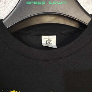 May include: Black crew neck t-shirt with a yellow brushstroke design. The shirt is hanging on a black hanger with the text "cropp town" in green. A tag with the letters "B&C COLLECTION" is visible.