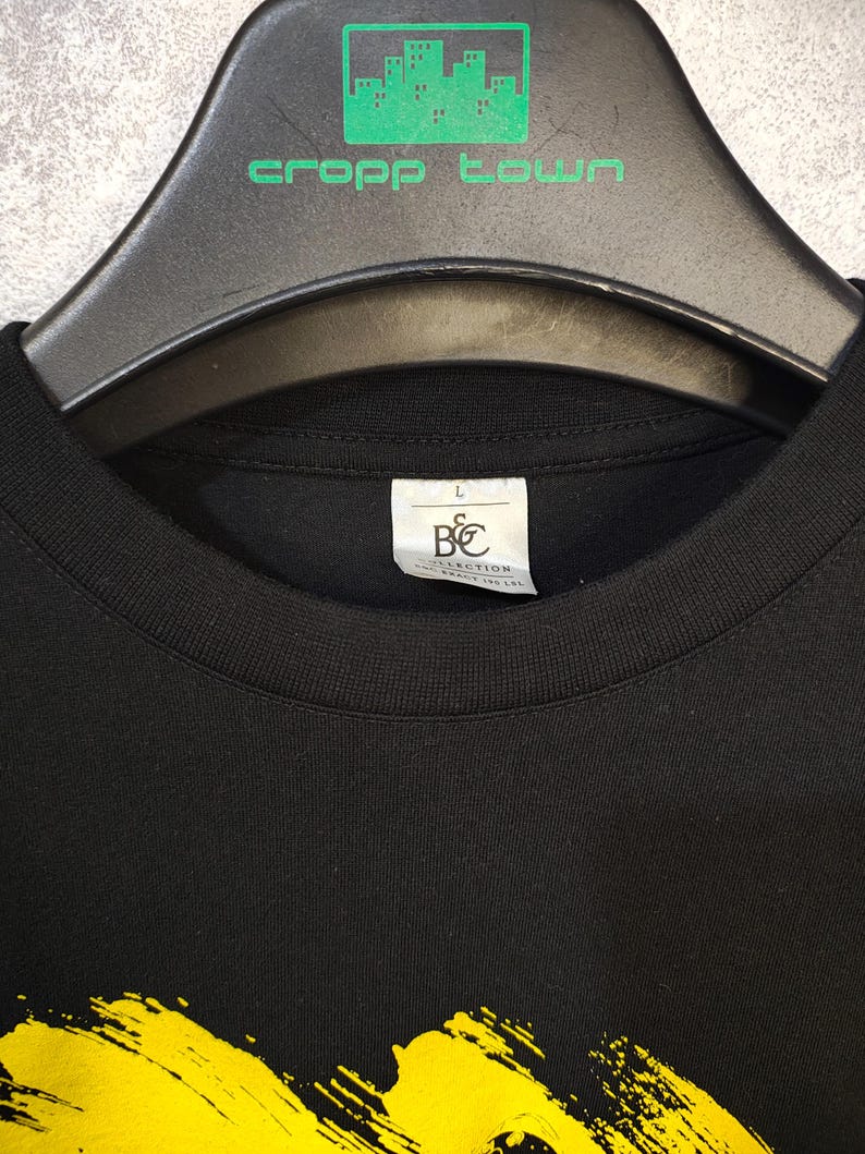 May include: Black crew neck t-shirt with a yellow brushstroke design. The shirt is hanging on a black hanger with the text "cropp town" in green. A tag with the letters "B&C COLLECTION" is visible.