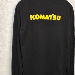 May include: Black long-sleeve shirt with the word "KOMATSU" printed in yellow across the back. The shirt is made of a soft material and has a crew neck. The shirt is hanging on a hanger against a gray background.