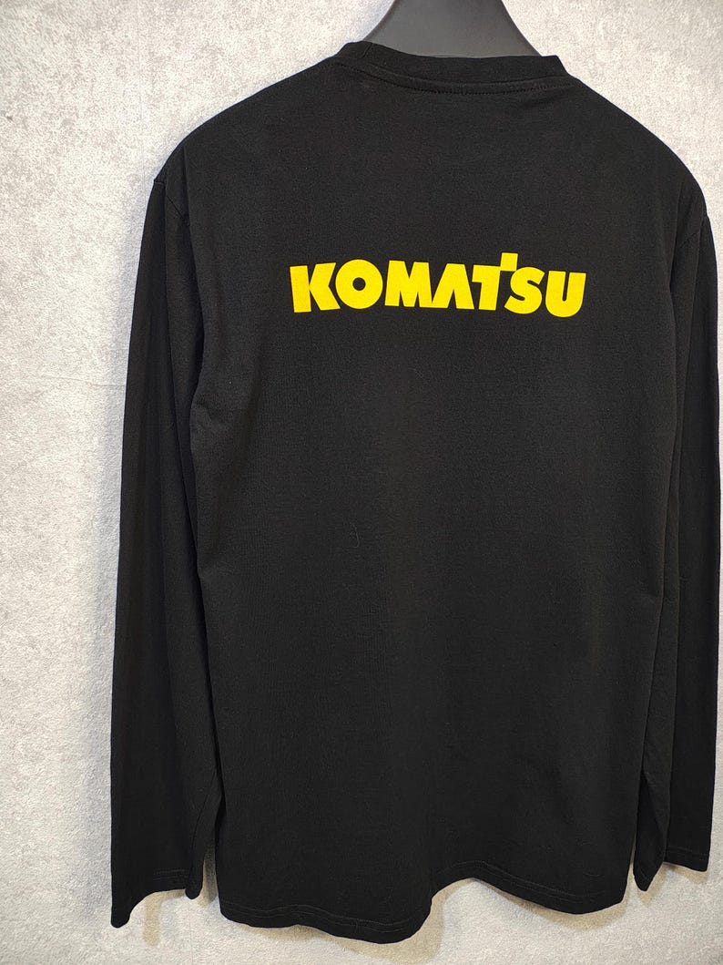 May include: Black long-sleeve shirt with the word "KOMATSU" printed in yellow across the back. The shirt is made of a soft material and has a crew neck. The shirt is hanging on a hanger against a gray background.