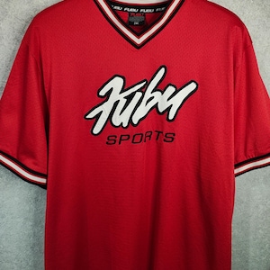 Op de afbeelding: Rood Fubu Sports shirt met een V-hals. Het shirt heeft het Fubu-logo in wit met een zwarte omlijning over de borst en het woord "SPORTS" eronder. De kraag en mouwboorden hebben zwart-witte strepen.