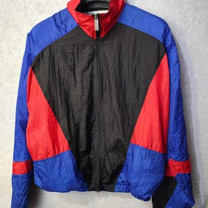 Può includere: Giacca retrò a blocchi di colore blu, rosso e nero. La giacca ha una cerniera completa, un colletto rialzato e polsini elasticizzati. Il design presenta un motivo geometrico con sezioni di colore audaci. La giacca è leggera e sembra essere realizzata in nylon o materiale simile.
