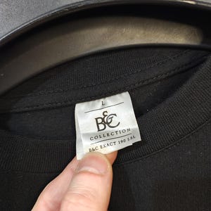 May include: Close-up of a black t-shirt with a white tag. The tag reads "L" and features the "B&C COLLECTION" logo and "B&C EXACT 190 LSL" text. The shirt is made of a dark fabric.
