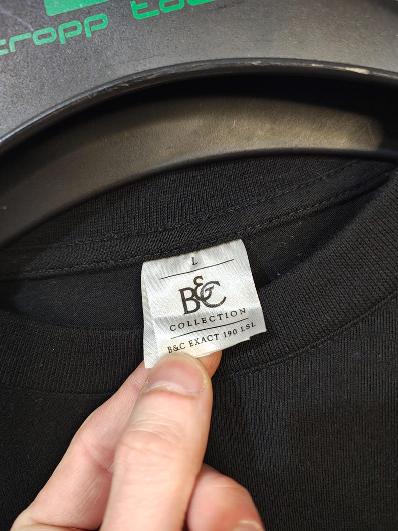 May include: Close-up of a black t-shirt with a white tag. The tag reads "L" and features the "B&C COLLECTION" logo and "B&C EXACT 190 LSL" text. The shirt is made of a dark fabric.