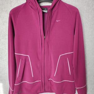 Puede incluir: Una sudadera con capucha magenta con cremallera, capucha y bolsillos. El logo de Nike está en la parte superior izquierda del pecho. Costuras blancas delinean los bolsillos y el dobladillo inferior. La palabra "Studio" está impresa en la parte inferior izquierda.