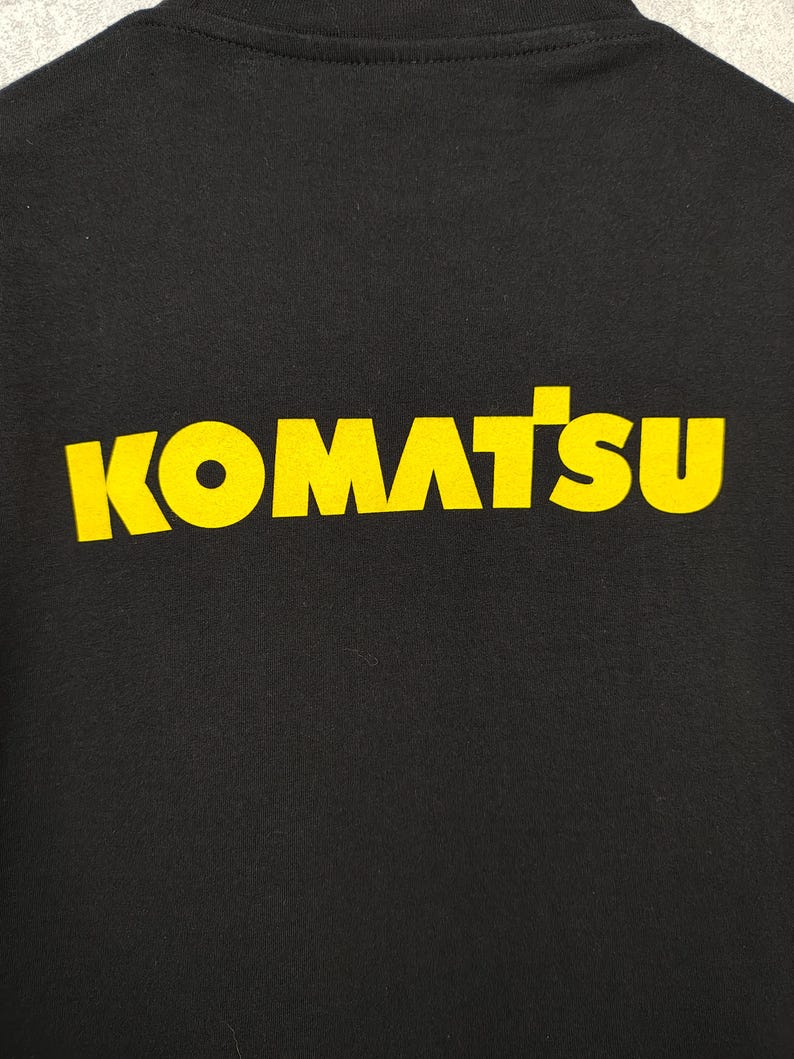 May include: Black t-shirt with the word "KOMATSU" printed in yellow block letters. The text is centered and prominently displayed on the front of the shirt. The shirt has a crew neck.