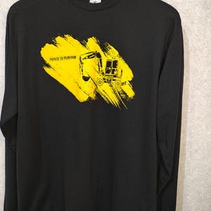 May include: Black long-sleeve shirt with a yellow graphic of a construction vehicle and the text "PROUD TO PERFORM". The graphic is on a yellow brushstroke background. The shirt is made of a soft material.