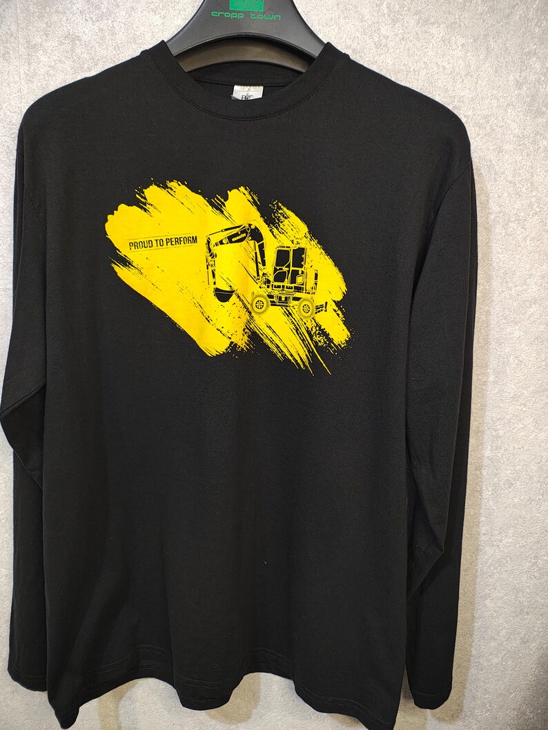 May include: Black long-sleeve shirt with a yellow graphic of a construction vehicle and the text "PROUD TO PERFORM". The graphic is on a yellow brushstroke background. The shirt is made of a soft material.