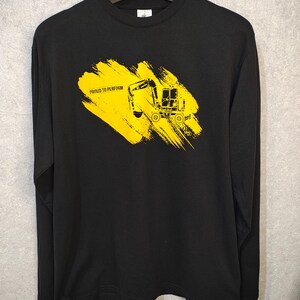 May include: Black long-sleeve shirt with a yellow graphic of a construction vehicle on a yellow brushstroke. The text "PROUD TO PERFORM" is above the vehicle. The shirt is hanging on a black hanger.