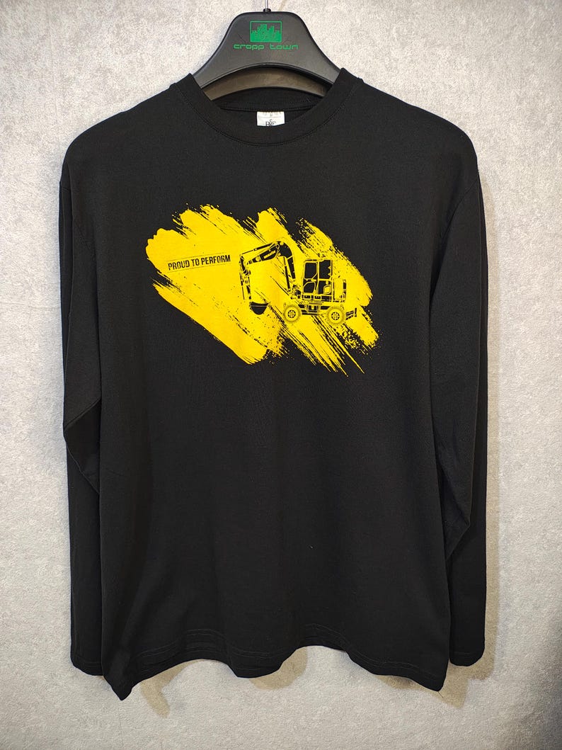 May include: Black long-sleeve shirt with a yellow graphic of a construction vehicle on a yellow brushstroke. The text "PROUD TO PERFORM" is above the vehicle. The shirt is hanging on a black hanger.