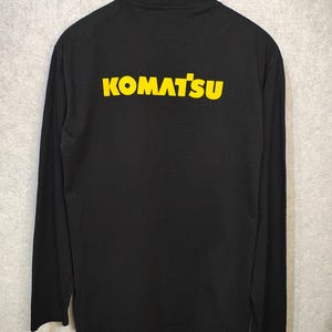 May include: Black long-sleeve shirt with the word "KOMATSU" printed in yellow across the back. The shirt is hanging on a black hanger against a light gray background. The text is in a bold, sans-serif font.