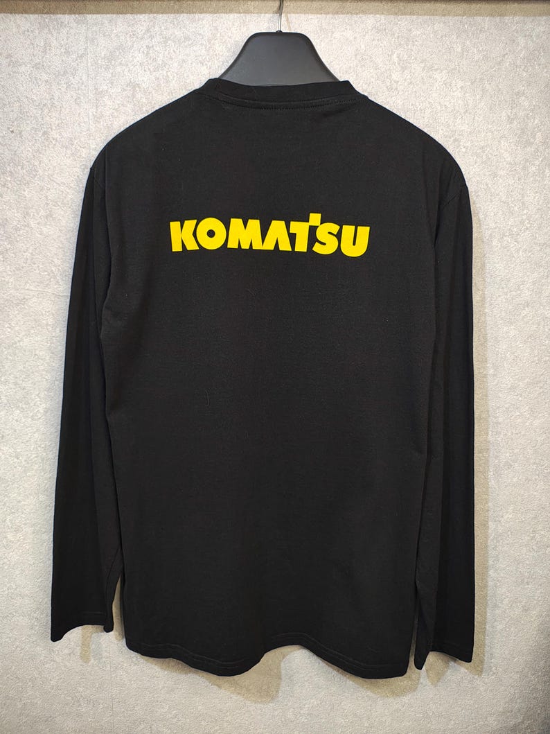 May include: Black long-sleeve shirt with the word "KOMATSU" printed in yellow across the back. The shirt is hanging on a black hanger against a light gray background. The text is in a bold, sans-serif font.