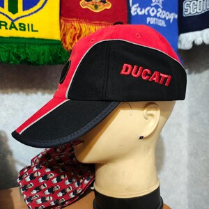 Op de afbeelding: Een rode en zwarte baseballcap met het woord "DUCATI" in het rood geborduurd. De cap heeft een gebogen klep met een zwart-witte streep. De cap wordt getoond op een mannequinhoofd.