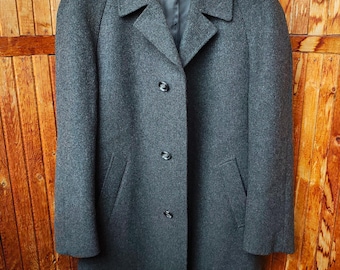 May include: A charcoal gray, knee-length overcoat with a classic design. The coat features a notched collar, three dark buttons, and two front pockets. It is hanging on a black hanger against a wooden background.