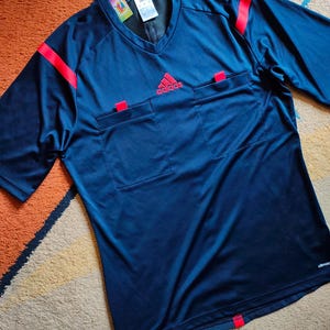 Puede incluir: Camiseta de árbitro Adidas azul marino con detalles rojos. Cuello en V, dos bolsillos en el pecho y rayas rojas en los hombros. El logo de Adidas es rojo. Fabricada en tejido transpirable.
