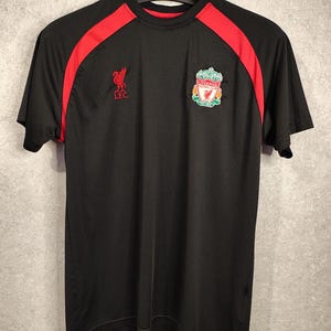 Può includere: Maglietta da calcio nera con inserti rossi sulle spalle. Presenta lo stemma del Liverpool Football Club e il logo L.F.C. La maglietta è appesa a una gruccia nera.