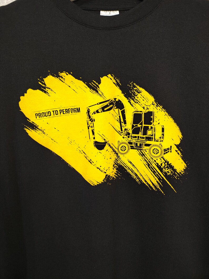 May include: Black sweatshirt featuring a yellow excavator graphic with the text "PROUD TO PERFORM" on a yellow brushstroke design. The excavator is a detailed illustration of heavy machinery.