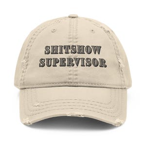 May include: A beige baseball cap with the text "SHITSHOW SUPERVISOR" in black, capital letters. The cap has a distressed, worn appearance, with a curved peak and visible wear and tear.