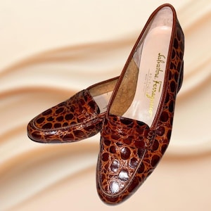 Puede incluir: Un par de mocasines de cuero marrón con estampado de cocodrilo. Los zapatos tienen un diseño clásico con punta redonda y tacón bajo. La marca "Salvatore Ferragamo" es visible en la plantilla.