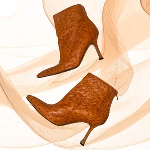 Puede incluir: Dos botines de cuero marrón con puntera afilada y tacones de aguja. Las botas tienen una superficie texturizada y una cremallera en la parte trasera. El fondo presenta un diseño suave y abstracto en tonos beige y marrón.